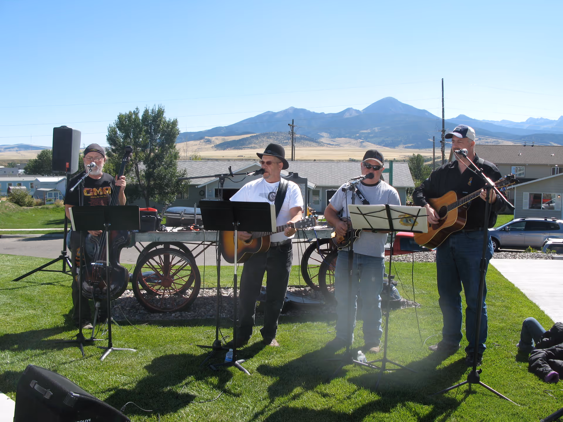 Four men playing musical instruments and singing outdoors on a grassy area with mountains and houses in the background. They have microphones and music stands in front of them, and there is a wagon wheel decoration behind them.
