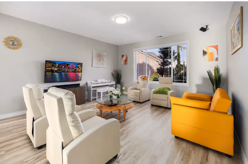 Bright living room with beige recliners, orange sofa, glass coffee table, TV, and large window.