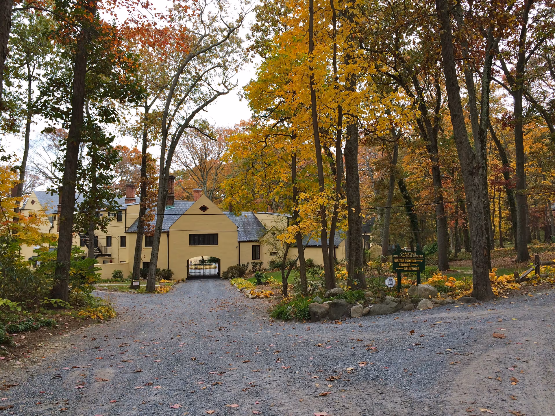 A gravel driveway leading to a yellow building with a gated archway, surrounded by tall trees with autumn foliage in shades of yellow and orange. There is a green sign near the driveway entrance that reads 'Christian Fellowship House' along with additional text. The scene is set in a wooded area with fallen leaves scattered on the ground.