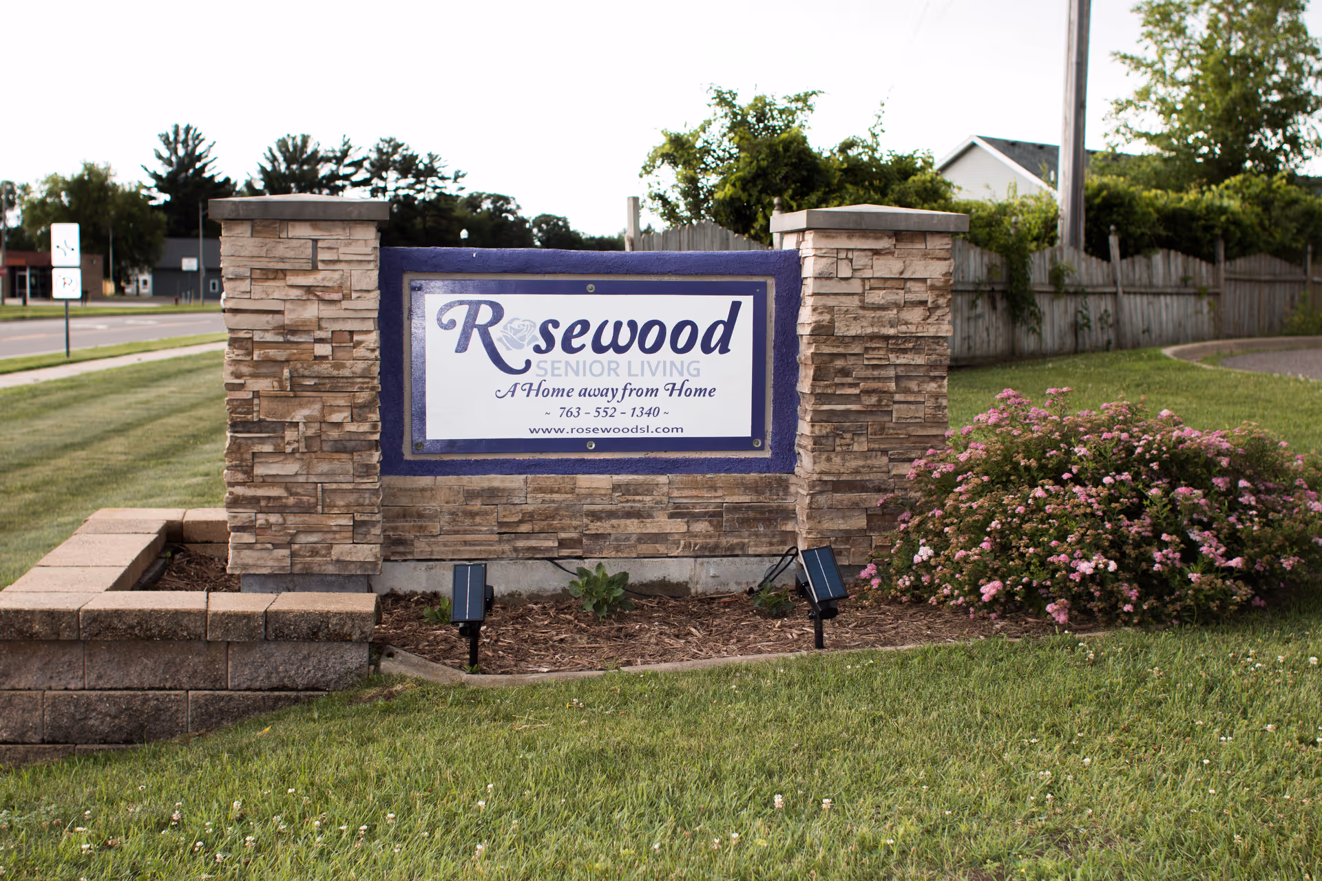 Stone sign for Rosewood Senior Living with the tagline 'A Home away from Home' and contact information, surrounded by grass and pink flowering bushes, with a road and trees in the background.