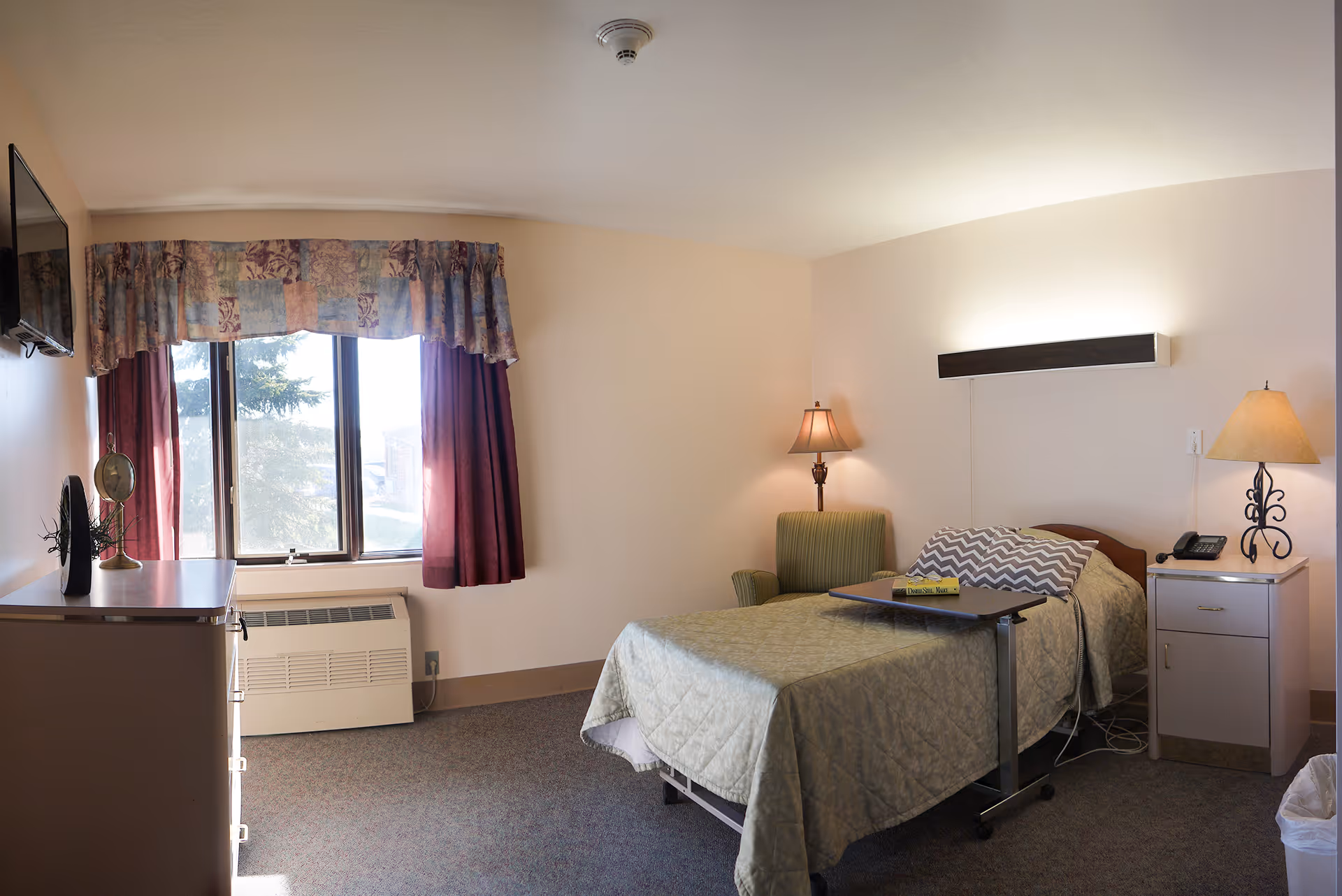 A single bedroom in a senior living facility with a bed covered in a green quilt, a chevron-patterned pillow, a small overbed table with a book, a green armchair, two lamps on either side of the bed, a window with floral and burgundy curtains, a wall-mounted TV, a dresser with a clock and plant, and a heating unit below the window.