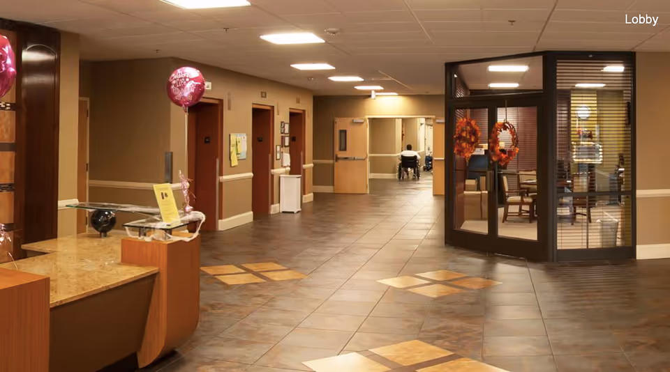 Interior view of a rehabilitation and wellness center lobby with a reception desk on the left, decorated with balloons. The hallway has several doorways and a person in a wheelchair is visible in the distance. There is a glass-enclosed office area on the right with autumn wreath decorations on the door.