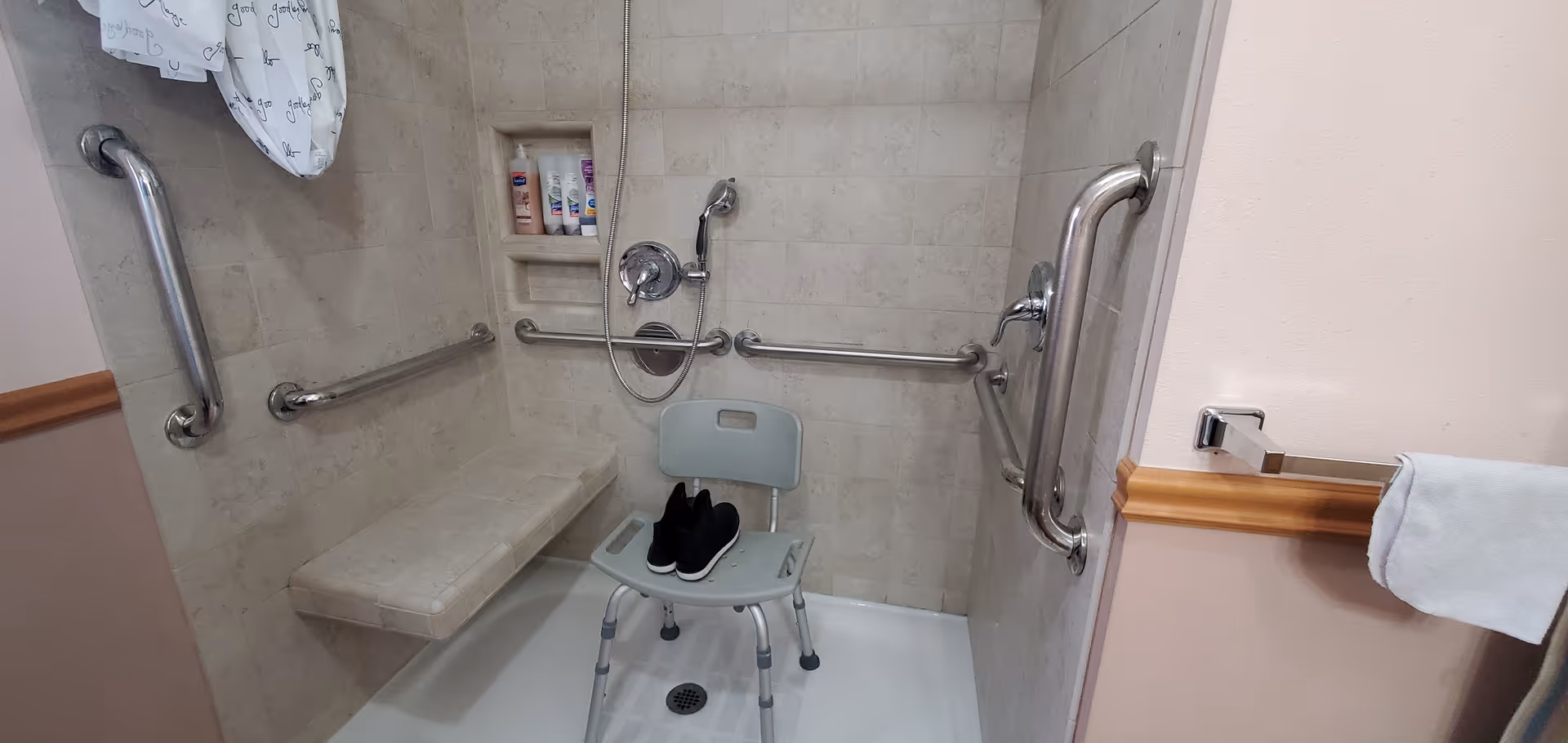 Accessible shower in a senior living facility with grab bars, a folding chair, and toiletries.