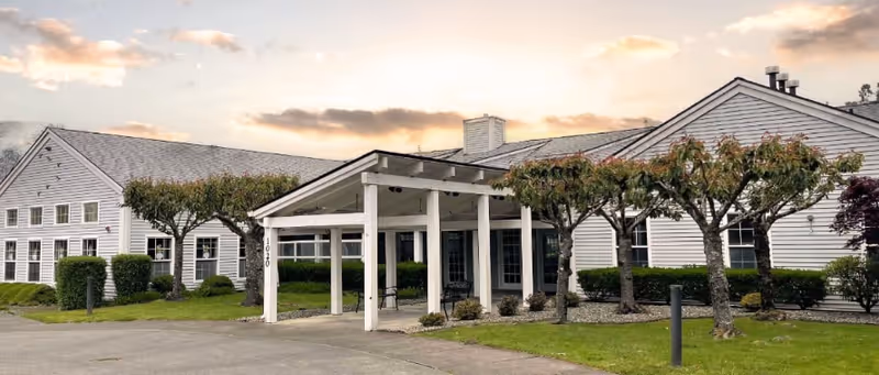 Exterior of a white senior living facility with a covered entrance and trimmed trees.