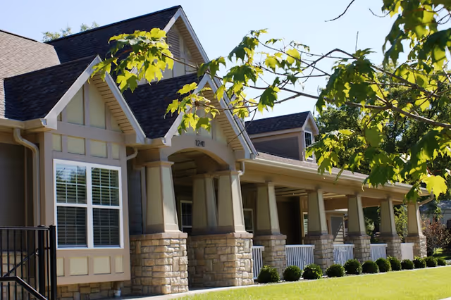 Senior living facility exterior with a covered porch, stone columns, and neatly trimmed lawn.