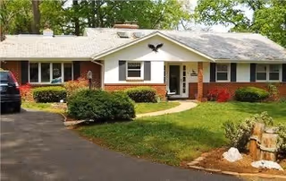Single-story ranch-style building with brick and white siding, a front lawn, driveway, shrubs, and surrounding trees.