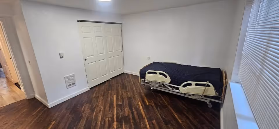 A small bedroom with a hospital-style adjustable bed, dark wood flooring, and white walls.