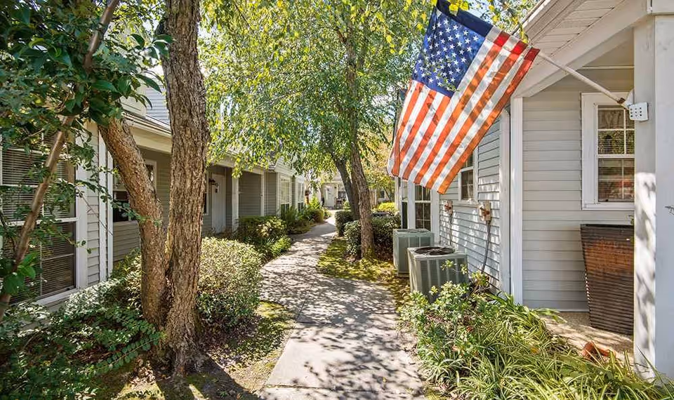 A sunny outdoor walkway between residential buildings with trees and shrubs lining the path. An American flag is mounted on the right building, and the buildings have light-colored siding with windows.