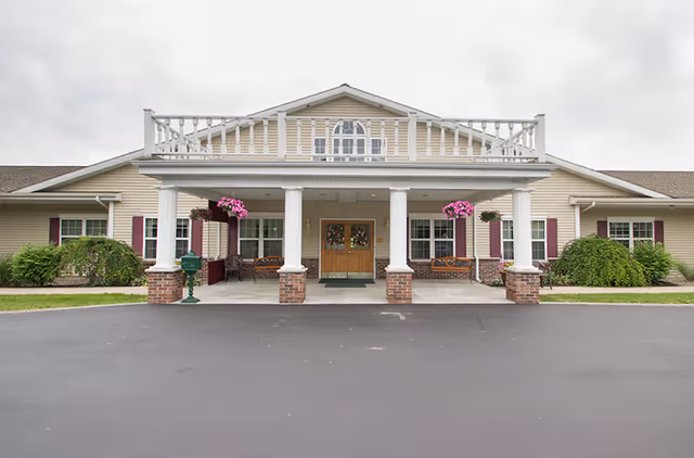 Front exterior view of a single-story assisted living community building with beige siding, white columns, and a covered entrance. There are hanging flower baskets and benches near the entrance, with a paved driveway in front.