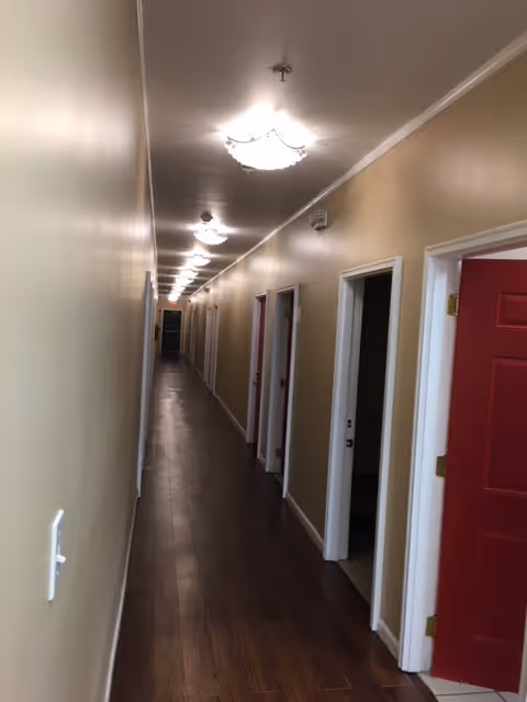 Long narrow hallway with wooden flooring and beige walls, multiple open doorways on the right side, ceiling lights evenly spaced along the corridor.