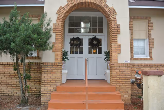 Entrance to a building with a brick archway and steps leading up to double white doors decorated with wreaths. There are potted plants on either side of the doors and a small tree to the left of the entrance.