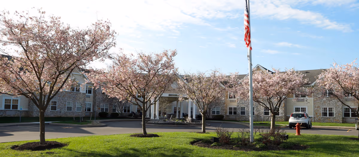 Exterior view of Clark Meadows facility with a circular driveway, blooming cherry blossom trees, an American flag on a flagpole, and a parked car near the entrance under a partly cloudy sky.
