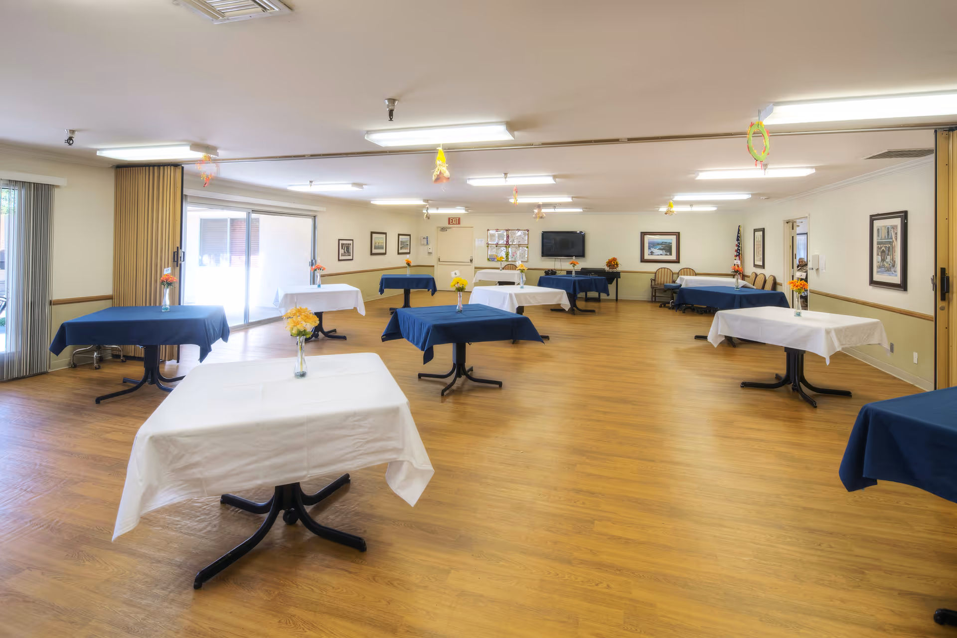 A spacious dining room with multiple tables covered in white and blue tablecloths, each adorned with a small vase of flowers. The room has wooden flooring, framed pictures on the walls, and large windows with vertical blinds allowing natural light to enter. There is a television mounted on the far wall and chairs lined up against the walls.