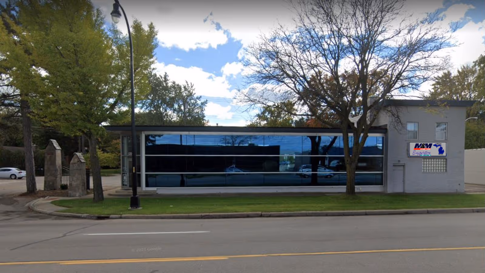 Glass-front facility building with large windows, trees, and a street view in front.