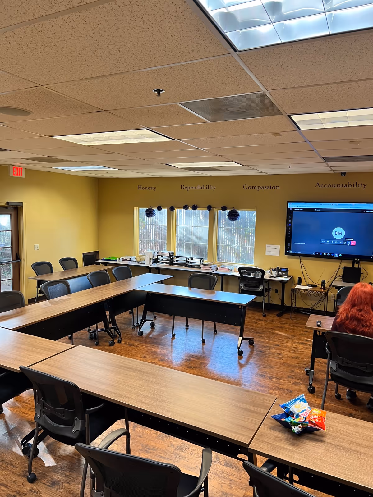 Classroom-style meeting room with tables, chairs, windows, wall slogans, and a large display screen.