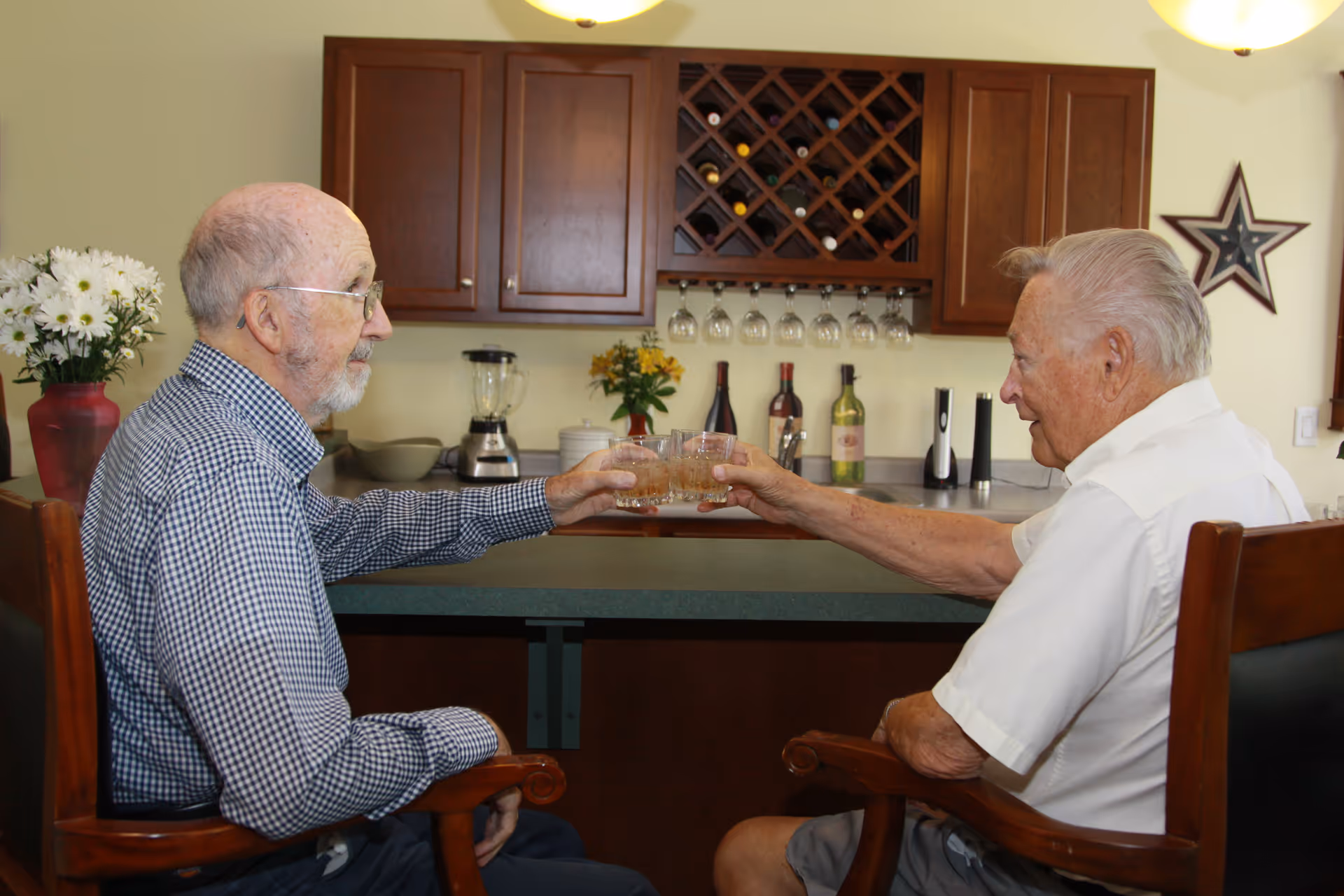 Two elderly men sitting on wooden chairs at a kitchen counter, clinking glasses in a toast. Behind them are wooden cabinets, a wine rack with bottles, hanging wine glasses, a blender, and some flowers in vases. A star-shaped wall decoration is visible on the right.