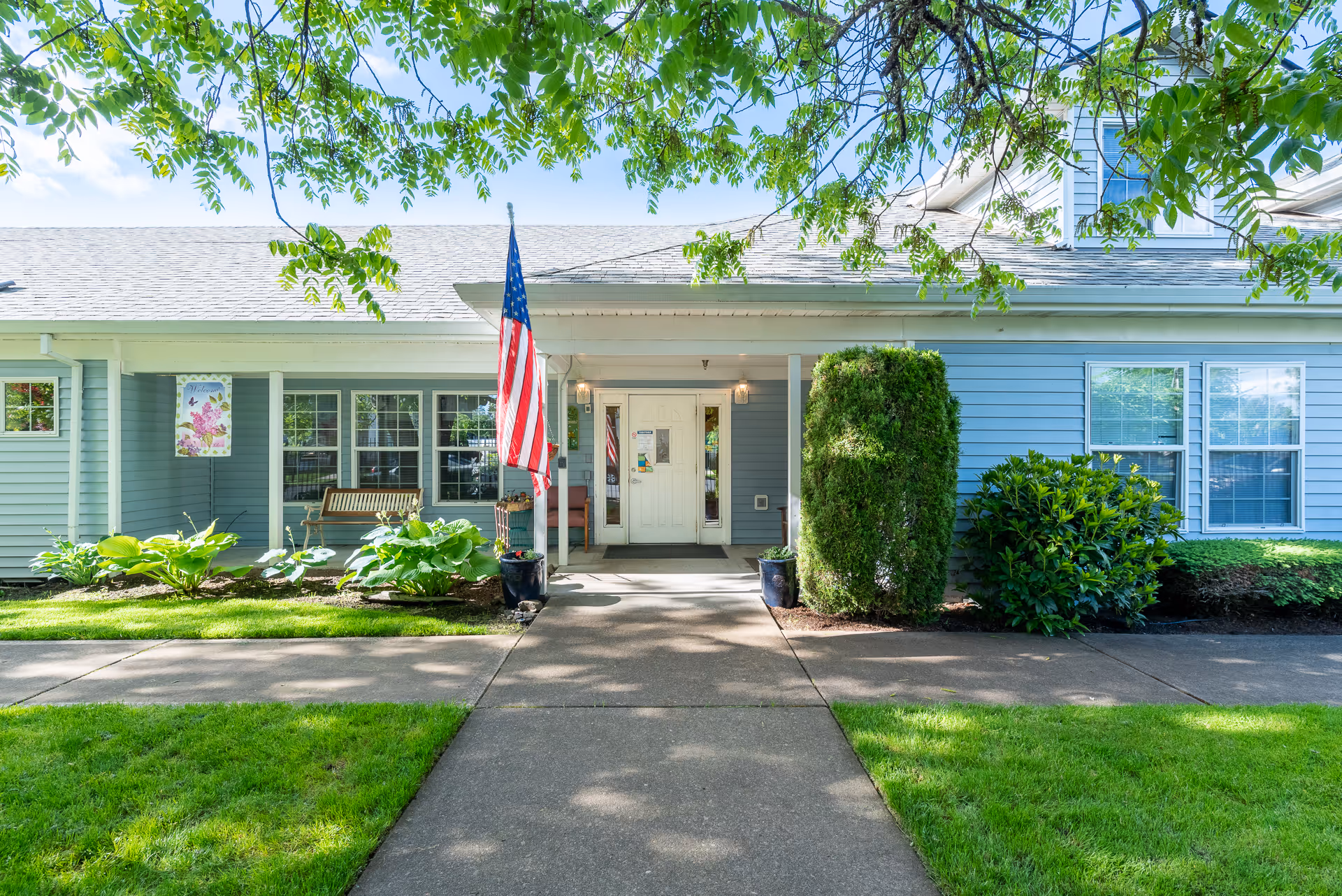 Front entrance of a light-blue single-story building with an American flag, bench, potted plants, and a paved walkway under leafy trees.