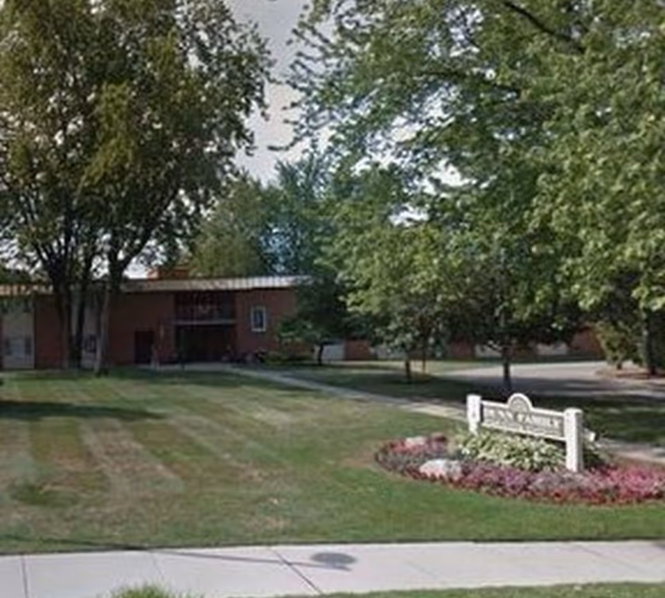 Brick senior apartment building set behind a grassy lawn and trees with a small landscaped sign near the sidewalk.