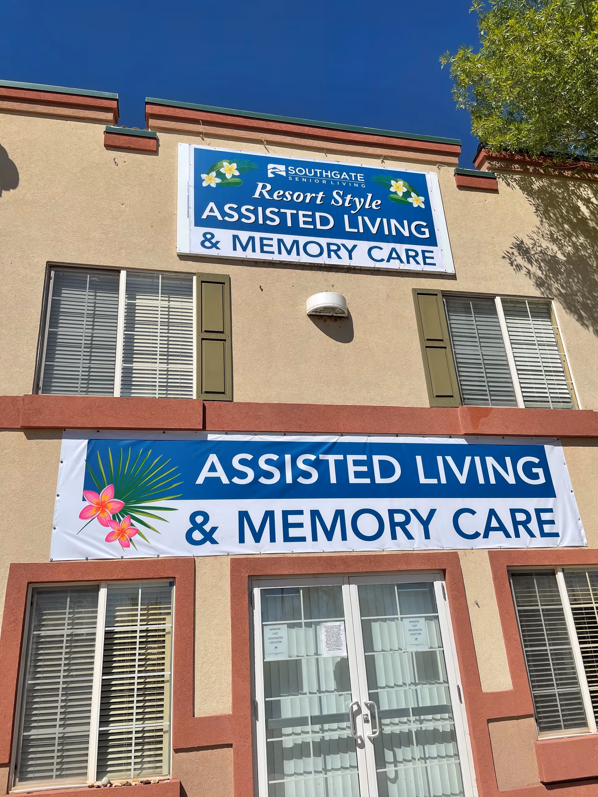 Exterior view of Southgate Senior Living facility showing two signs that read 'Resort Style Assisted Living & Memory Care' on a beige building with red trim, windows with shutters, and glass doors under a clear blue sky.