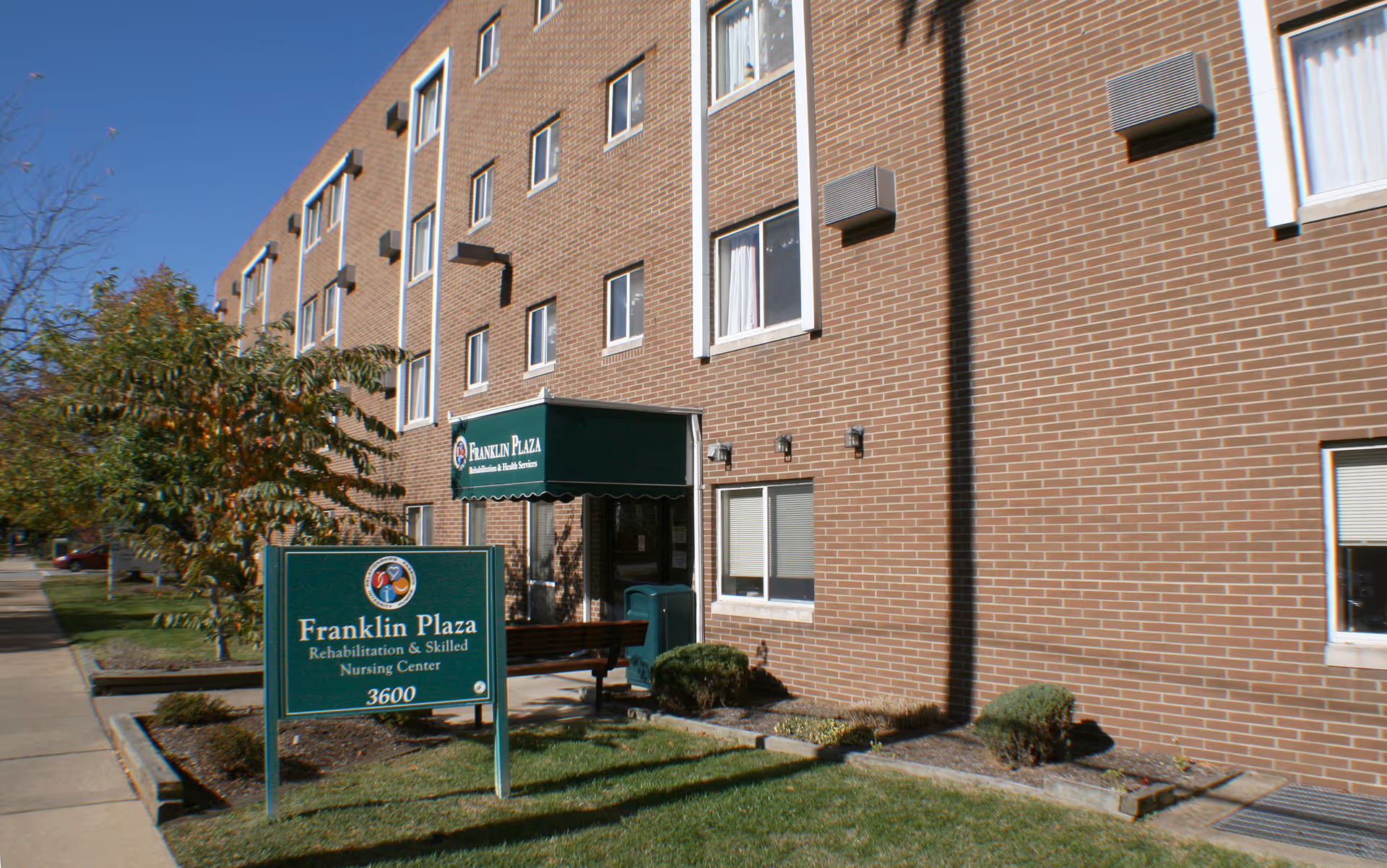 Exterior view of Franklin Plaza Rehabilitation & Skilled Nursing Center, a multi-story brick building with several windows, a green awning over the entrance, and a green sign in front displaying the facility name and address 3600. There is a sidewalk and some landscaping with bushes and a tree.