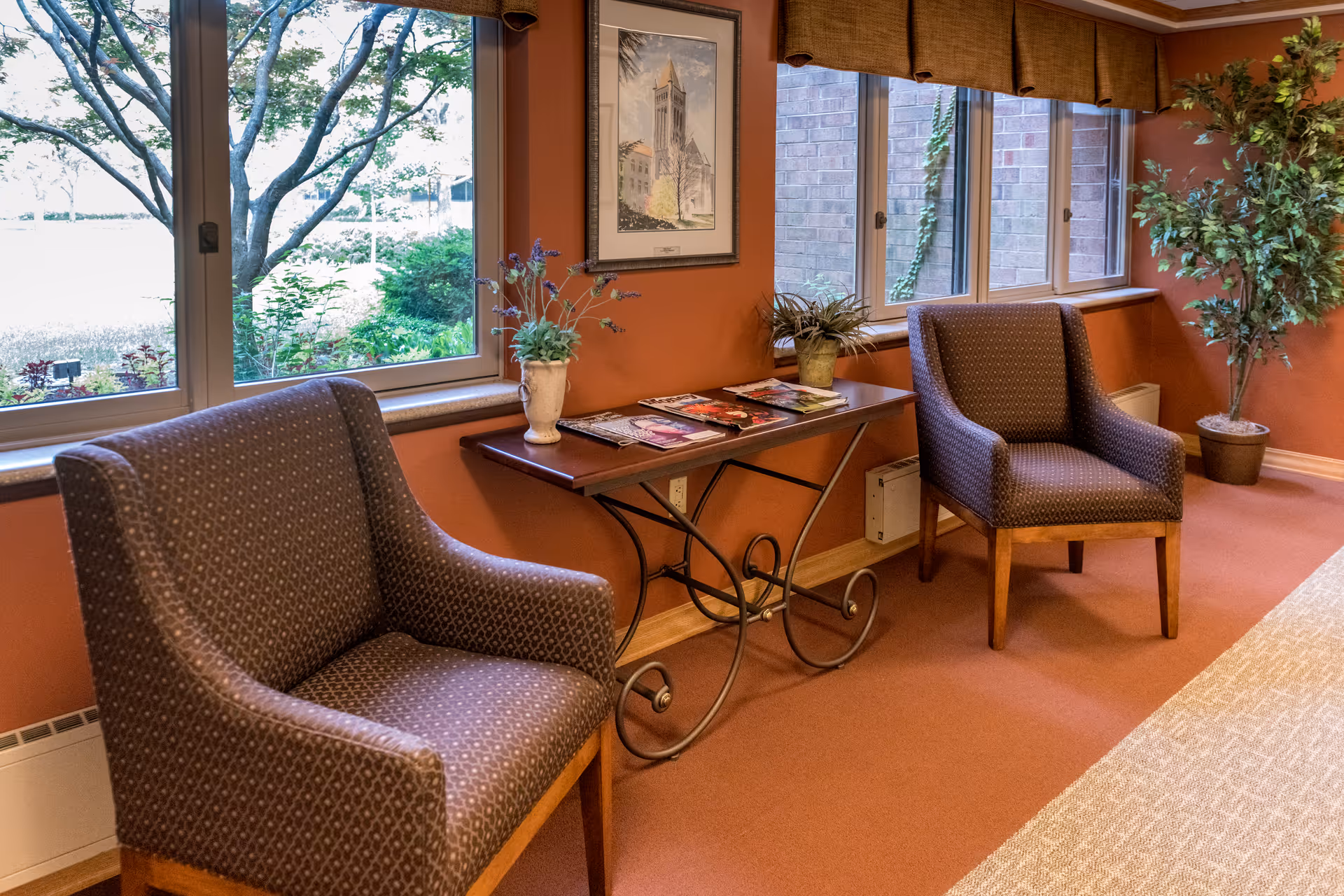Two upholstered chairs flank a decorative console table with magazines beneath windows looking out onto greenery.