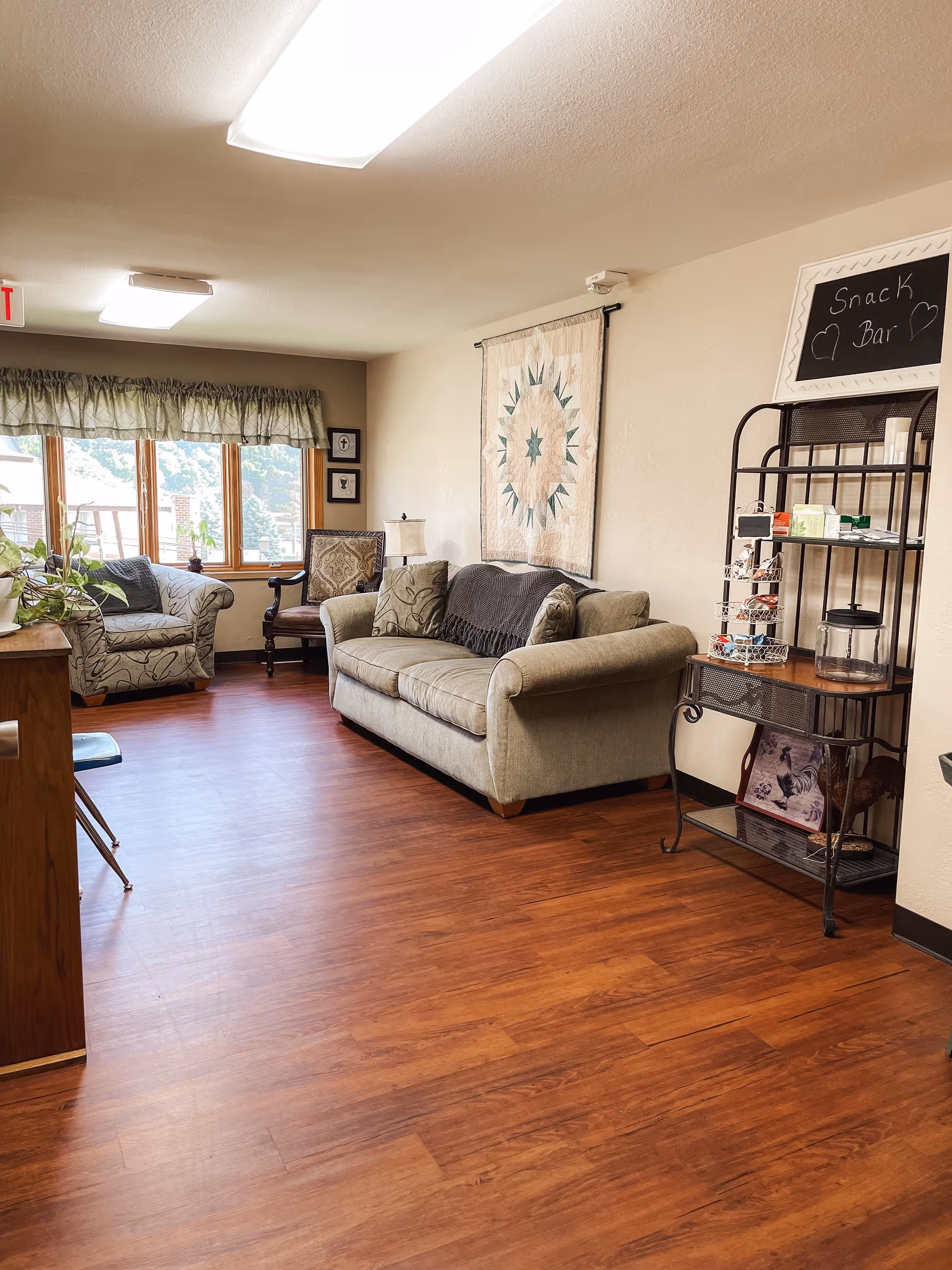 A cozy common area with wooden flooring, featuring a beige sofa with cushions, an armchair, and a wooden chair near a window with green curtains. On the wall above the sofa is a decorative quilt. To the right, there is a metal snack bar rack with various items and a small chalkboard sign that reads 'Snack Bar' with hearts drawn on it.