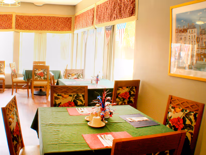 Dining room with several tables covered in green tablecloths, each set with placemats, utensils, and cups. Chairs have floral patterned cushions. Large windows with red valances let in natural light. A framed picture hangs on the wall.