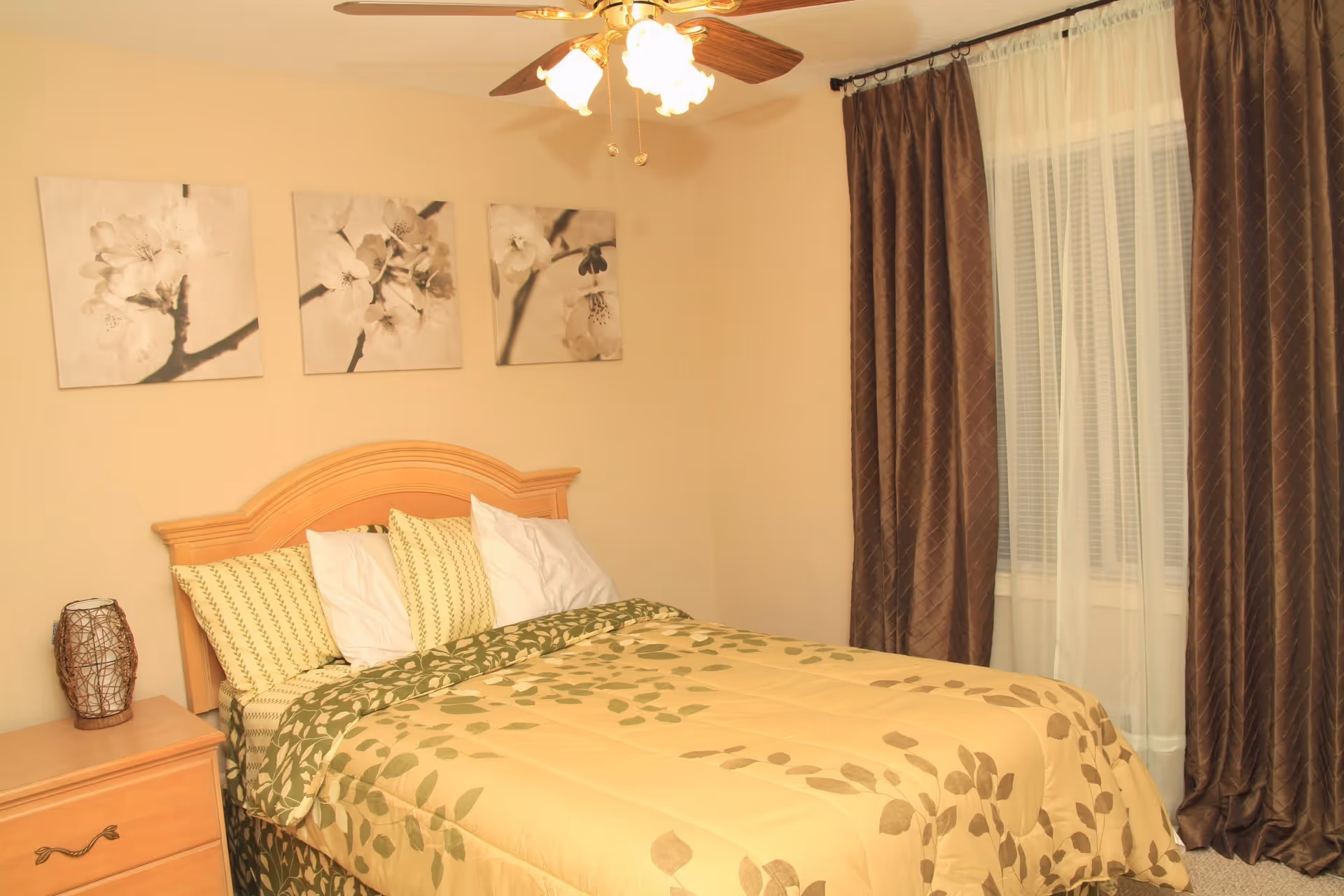 A cozy bedroom with a wooden headboard, patterned bedding, nightstand, curtains, and a ceiling fan.