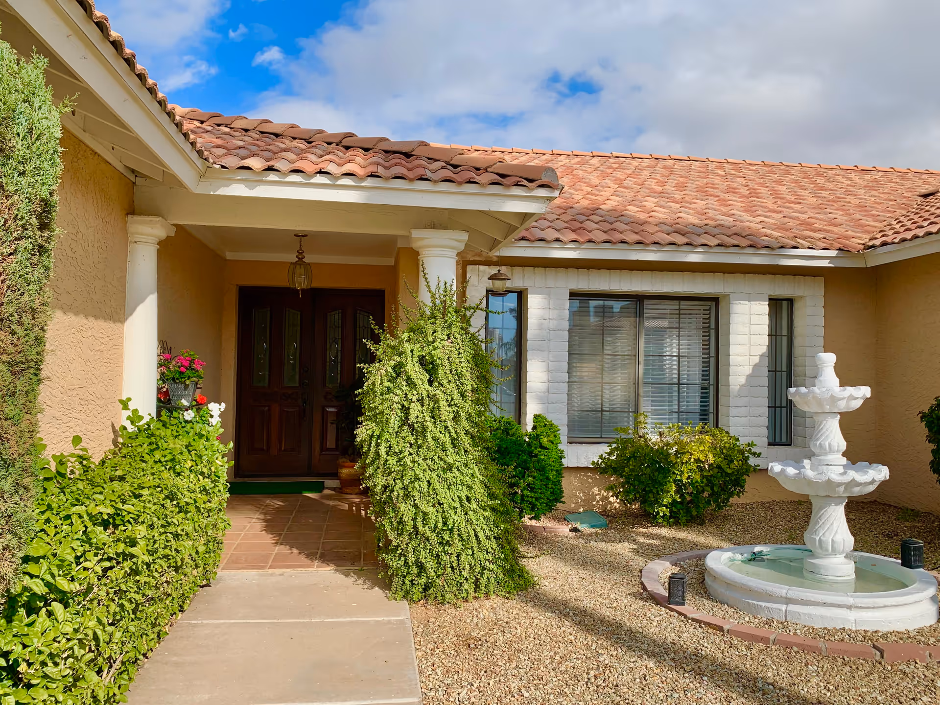 Front entrance of a single-story building with a tiled roof, beige stucco walls, and a covered porch supported by white columns. There is a dark wooden double door at the entrance, surrounded by green bushes and plants. To the right, there is a white two-tiered water fountain on a gravel area with some small shrubs and a window with white trim and blinds.