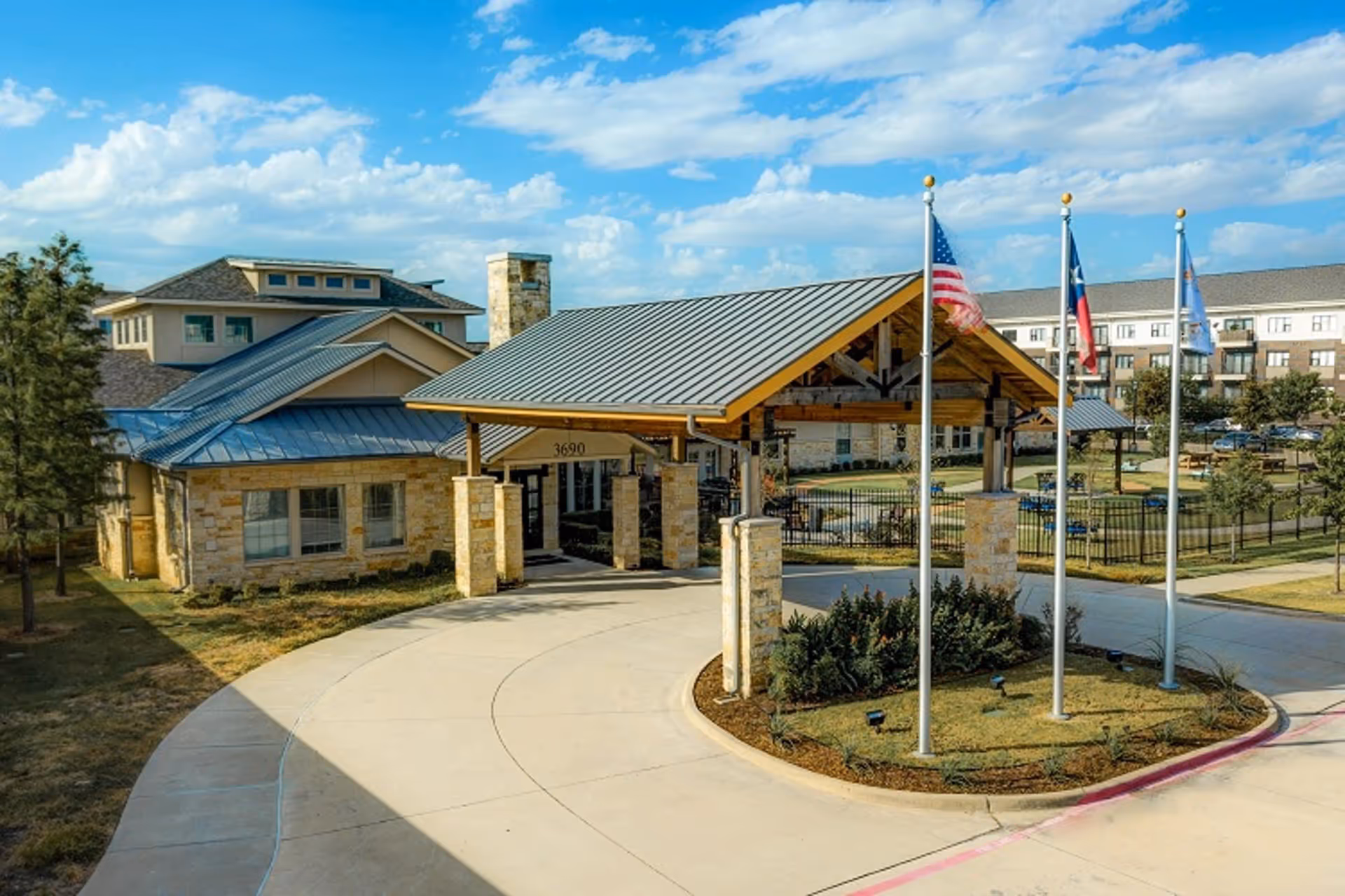 Covered entrance canopy and circular driveway of a senior living facility with flagpoles and surrounding buildings under a blue sky.