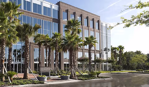 Modern glass-front senior living building with palm trees and a sunny parking lot