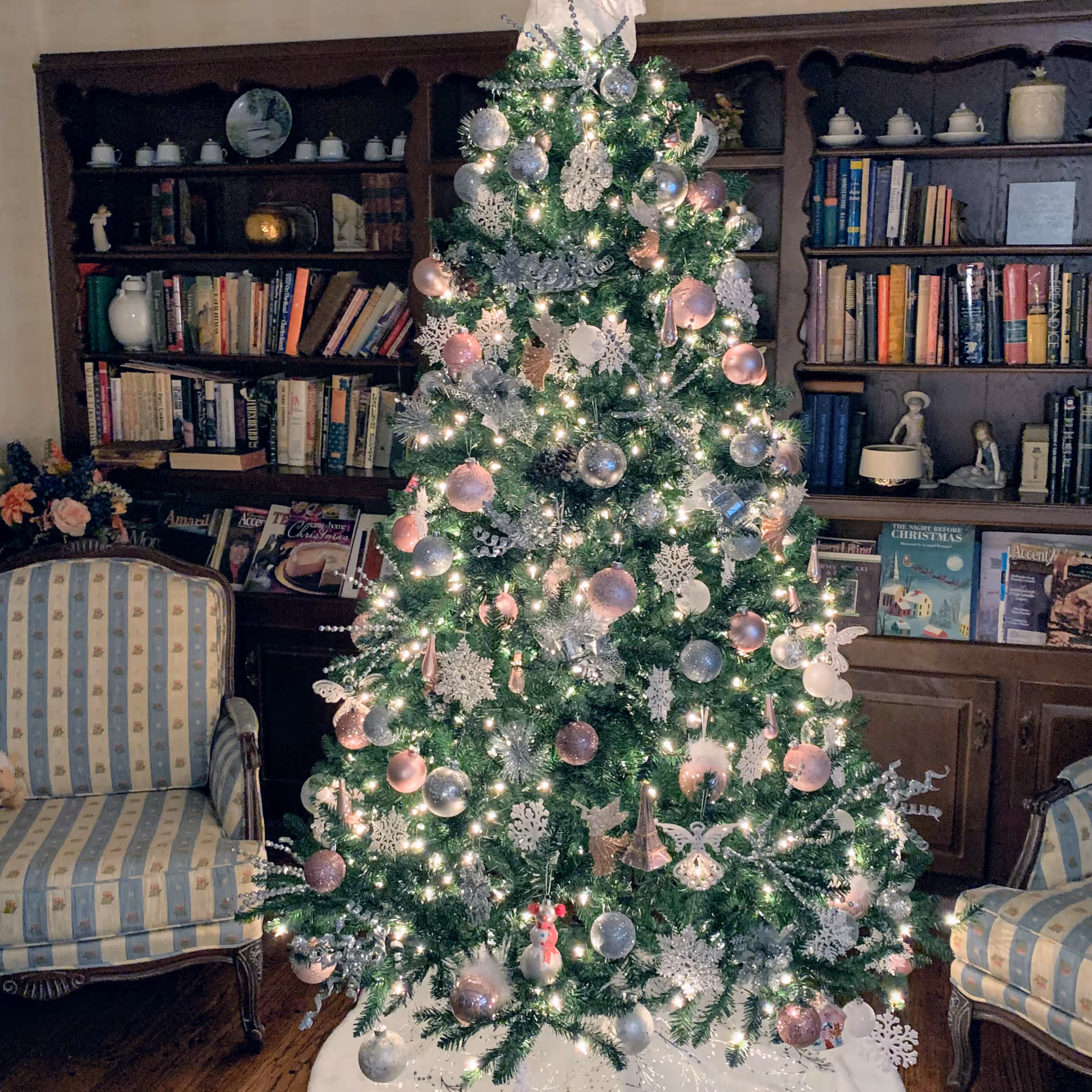 Decorated Christmas tree in cozy living room with bookshelves and upholstered chairs.