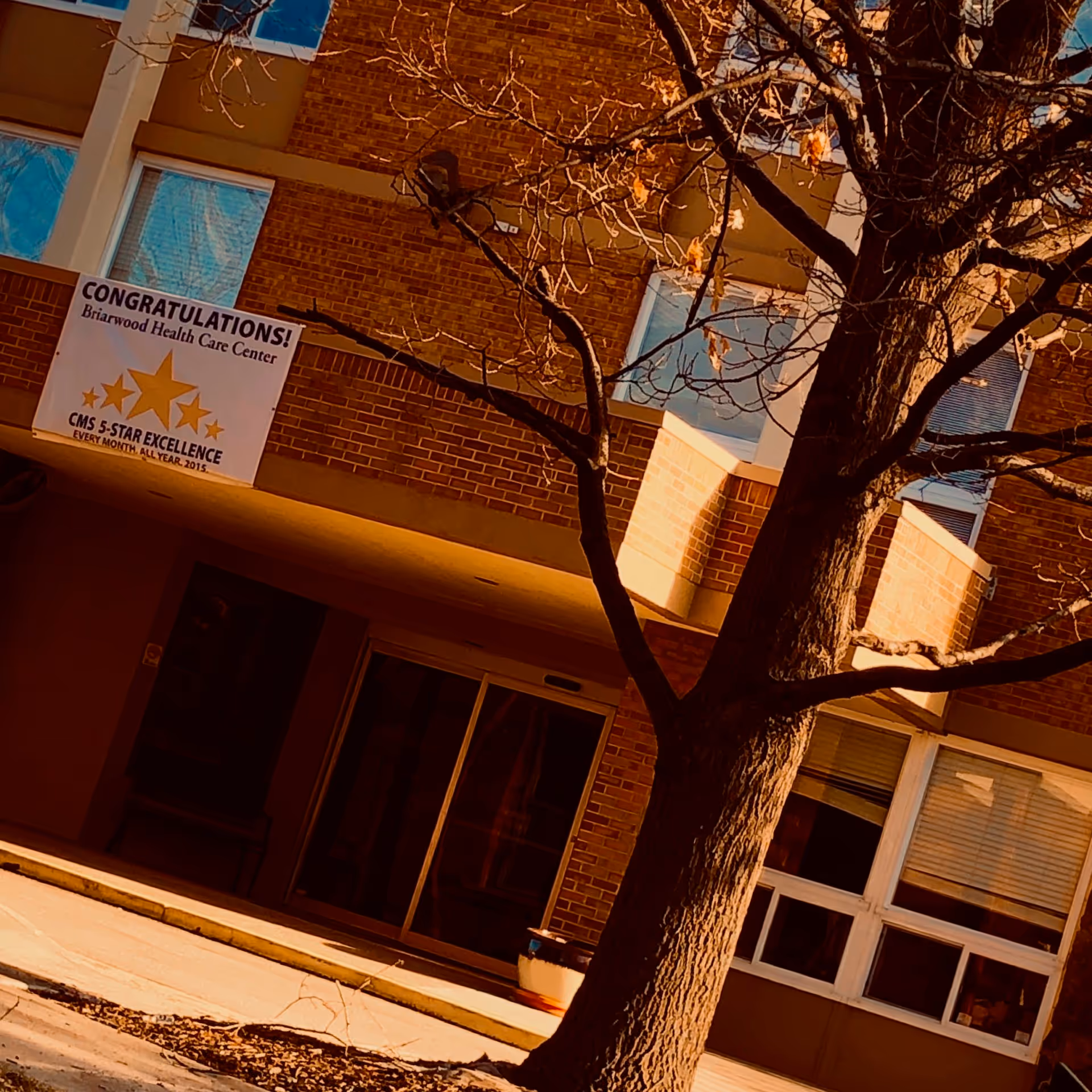 Exterior view of Briarwood Health Care Center with a banner and a tree in foreground