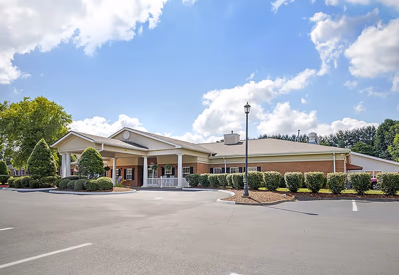 Front entrance of a one-story brick senior living facility with a covered portico, parking lot and landscaped shrubs under a blue sky.