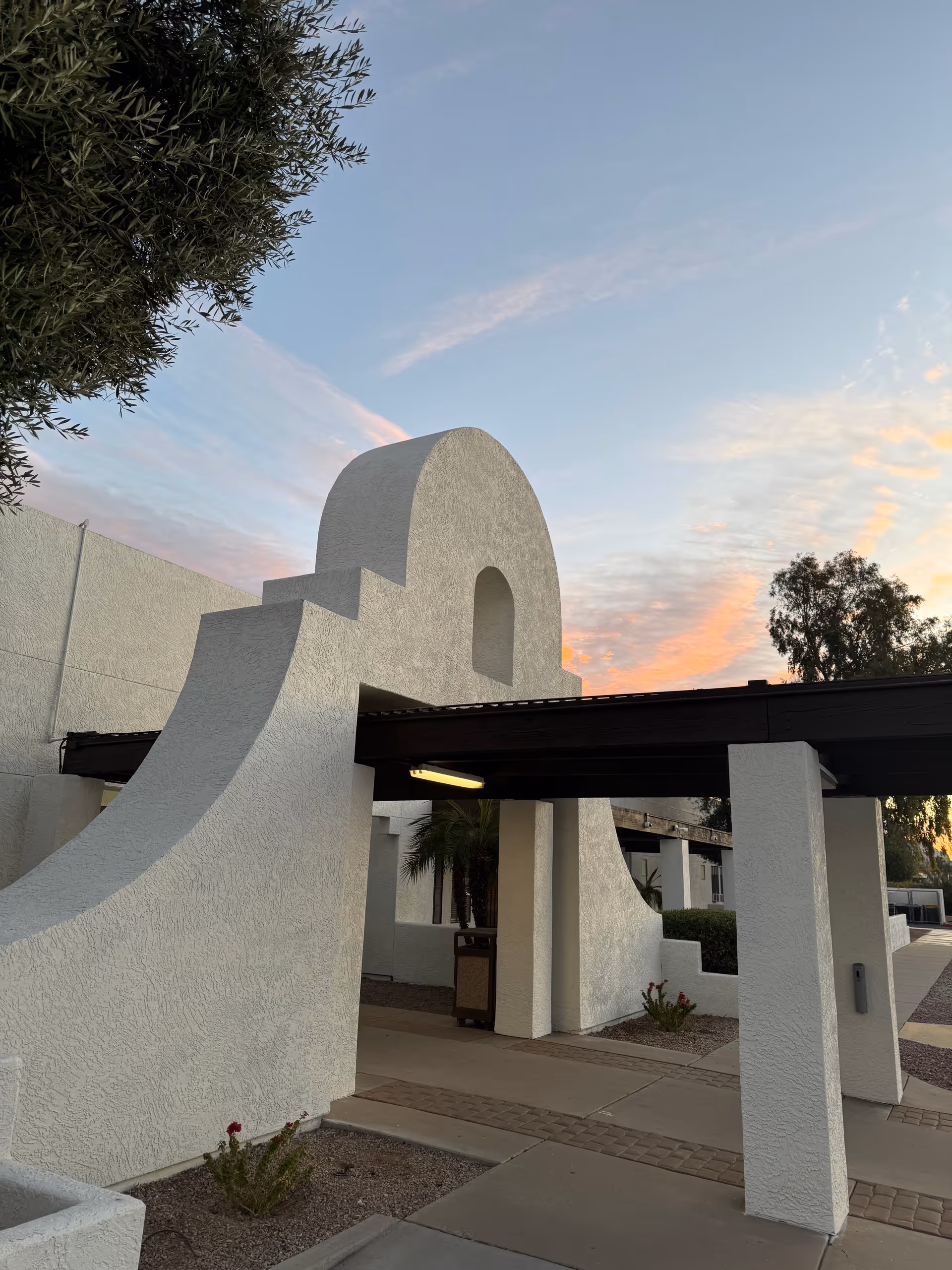 White stucco arches and a covered walkway against a sunset sky at the facility entrance.
