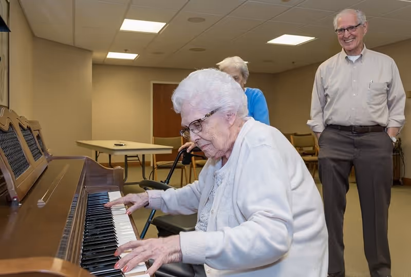 An elderly woman with white hair and glasses is playing a wooden piano in a room. Behind her, another elderly woman with white hair and a blue sweater is watching, and an elderly man wearing glasses, a beige shirt, and dark pants is smiling while observing the piano player. The room has beige walls, a table, and chairs in the background.