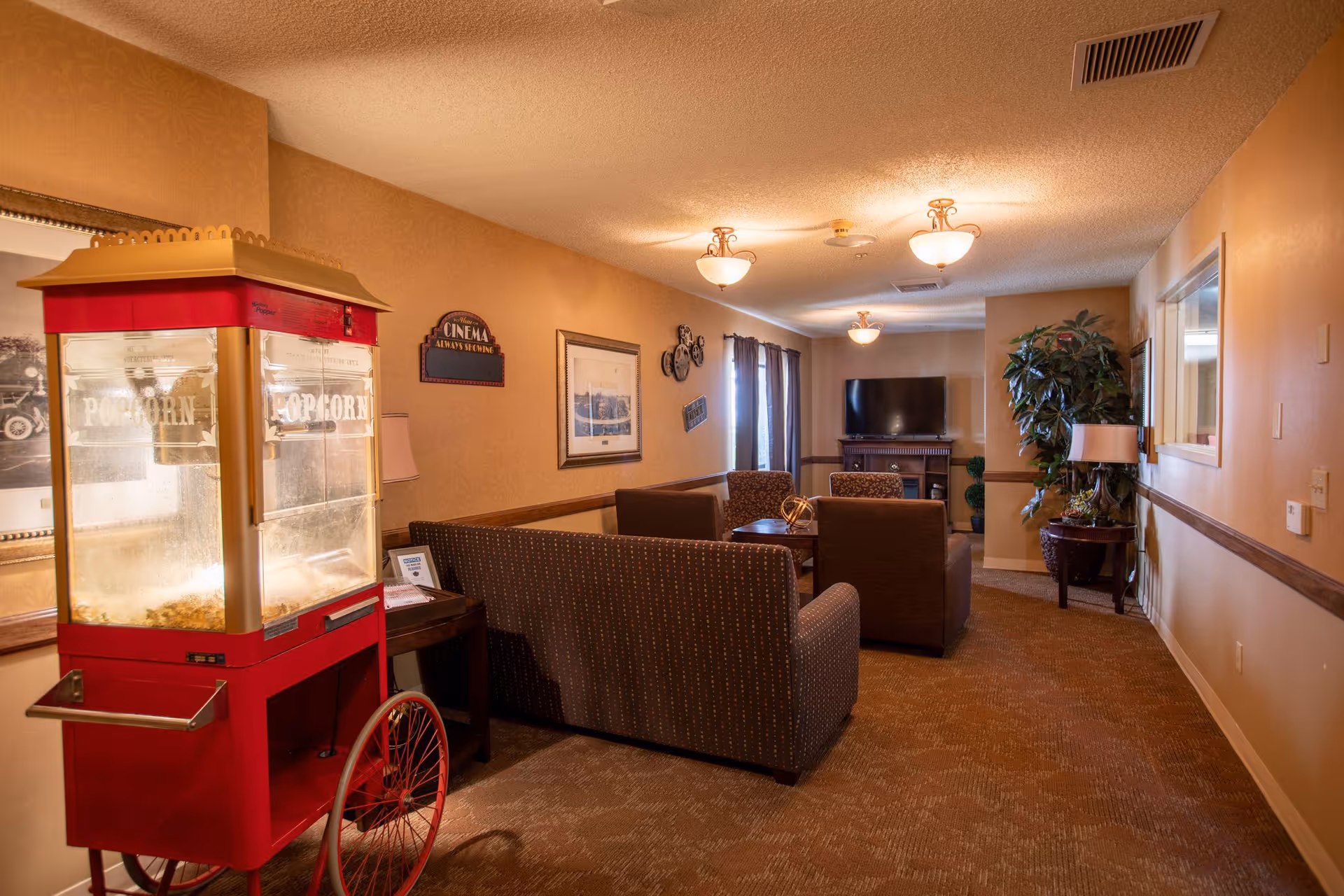 A cozy senior living common area with a red popcorn machine on the left, several brown upholstered sofas and chairs arranged around small tables, a flat-screen TV mounted on a stand at the far end, warm lighting from ceiling fixtures, framed pictures and decorative signs on the walls, and a large potted plant near the TV.