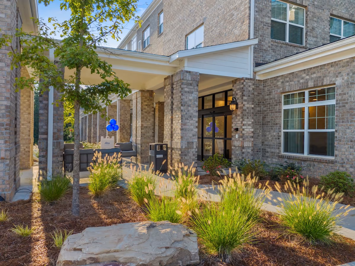 Brick-facade senior living facility entrance with glass doors and landscaped walkway.
