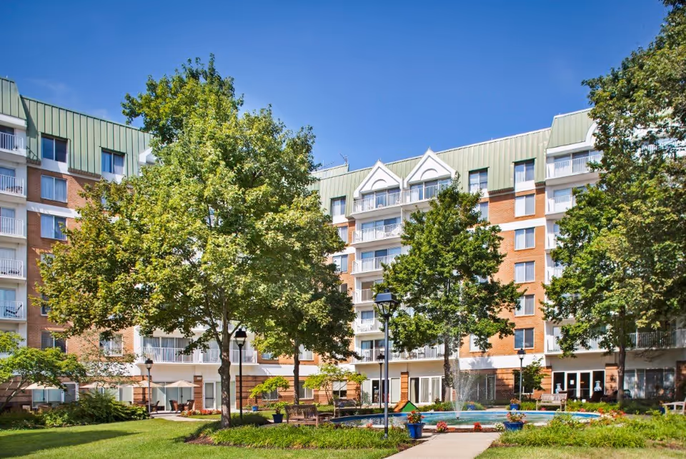 Exterior view of Bedford Court senior living facility with a multi-story brick building featuring balconies. In the foreground, there is a landscaped garden area with green trees, a walkway, benches, and a circular water fountain under a clear blue sky.