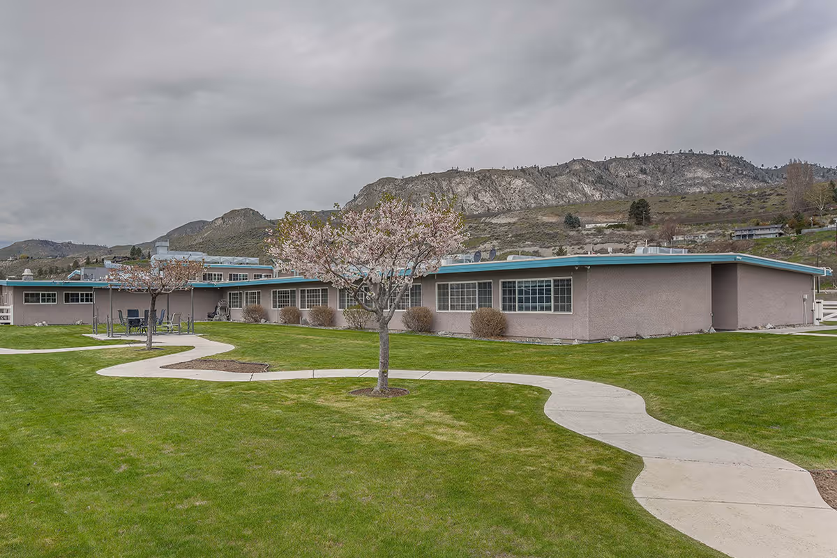 Single-story beige rehab facility with curved walkway, blooming trees, and mountain backdrop.