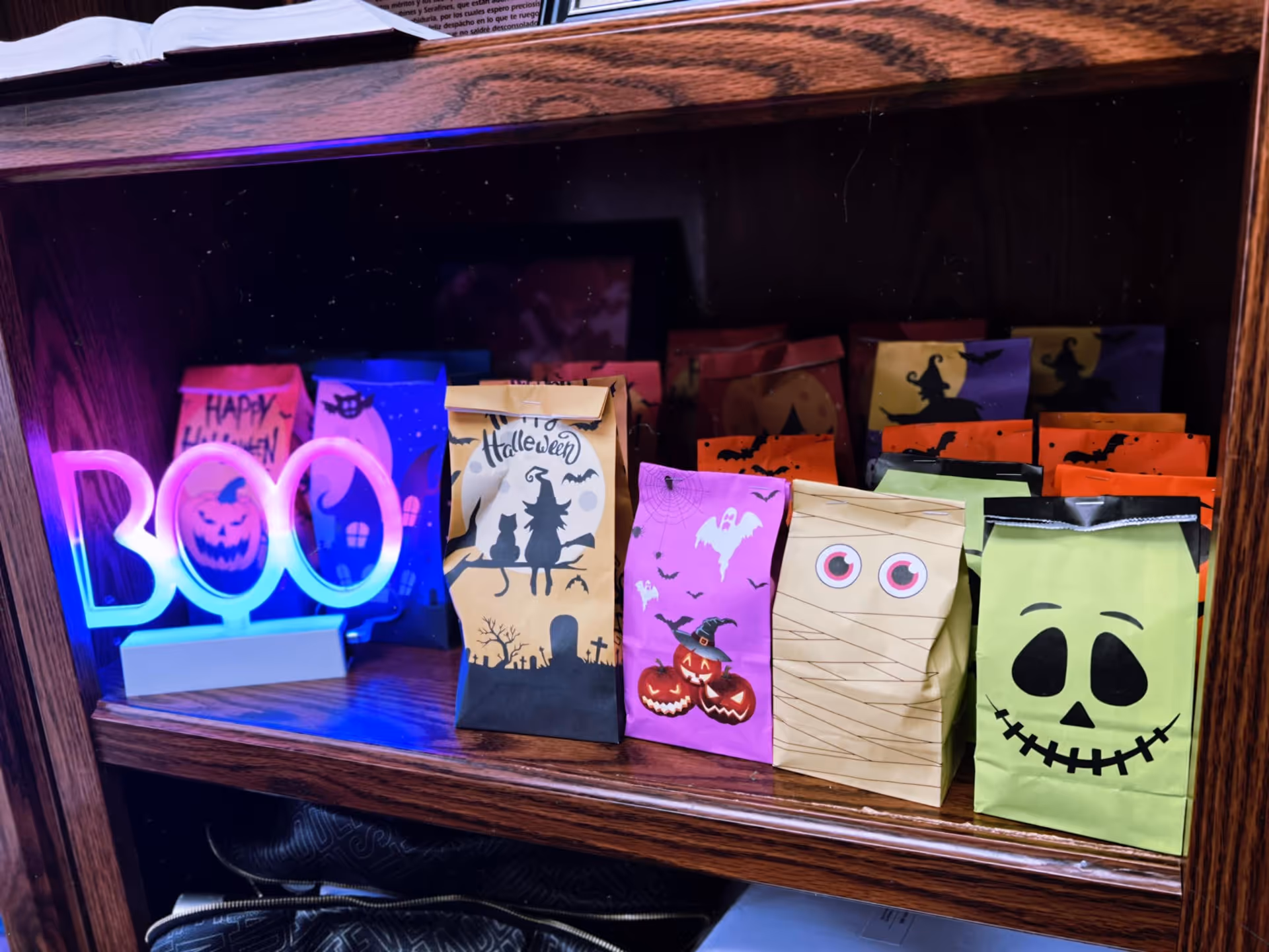 Halloween gift bags on a wooden shelf beside a neon 'BOO' sign