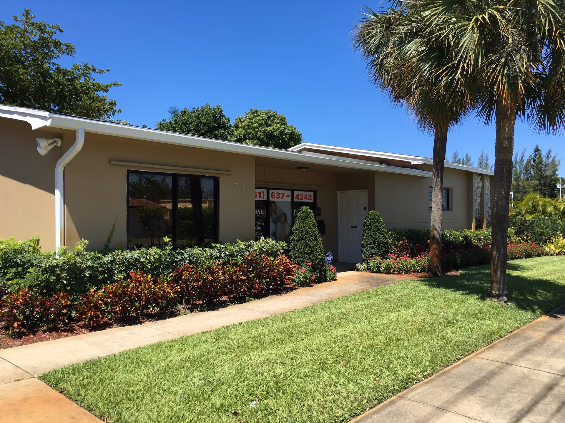 Single-story beige senior care facility with large windows, landscaped front yard, and palm trees.