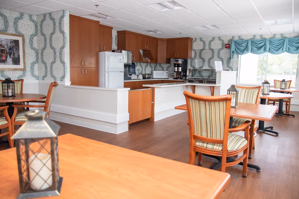 Dining area in a senior living facility with wood tables and a back kitchenette.