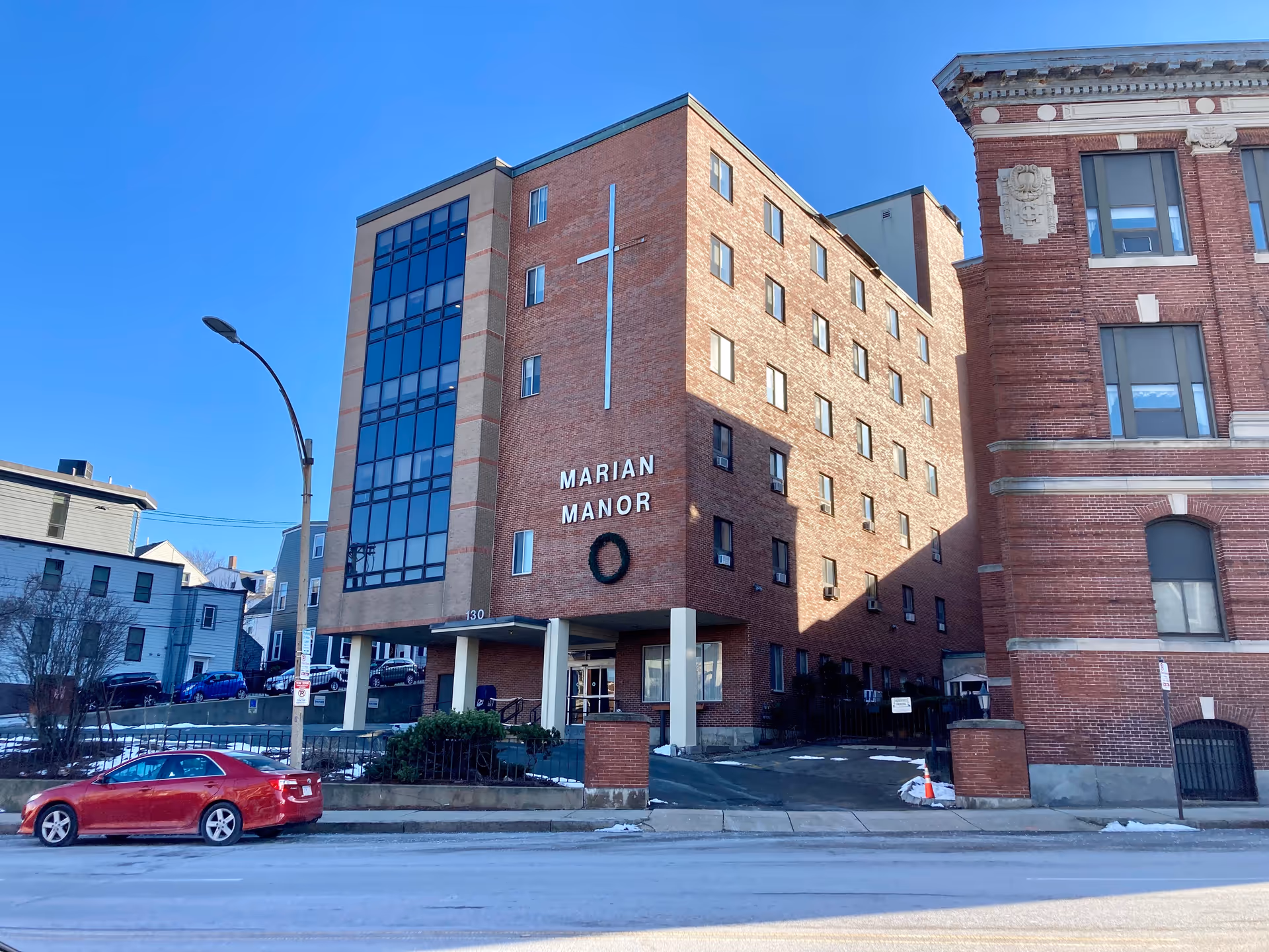 Front exterior of a multi-story brick building labeled "Marian Manor" with a cross on the facade and a red car parked in front.