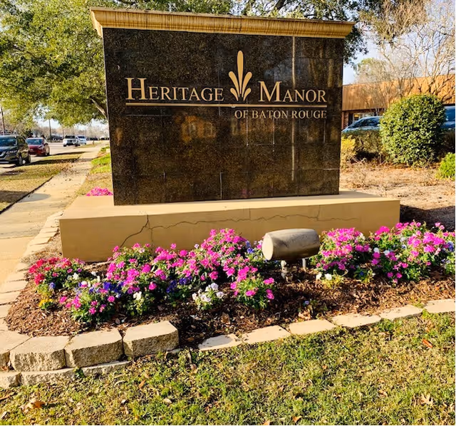 A large black granite sign for Heritage Manor of Baton Rouge sits among blooming flowers.