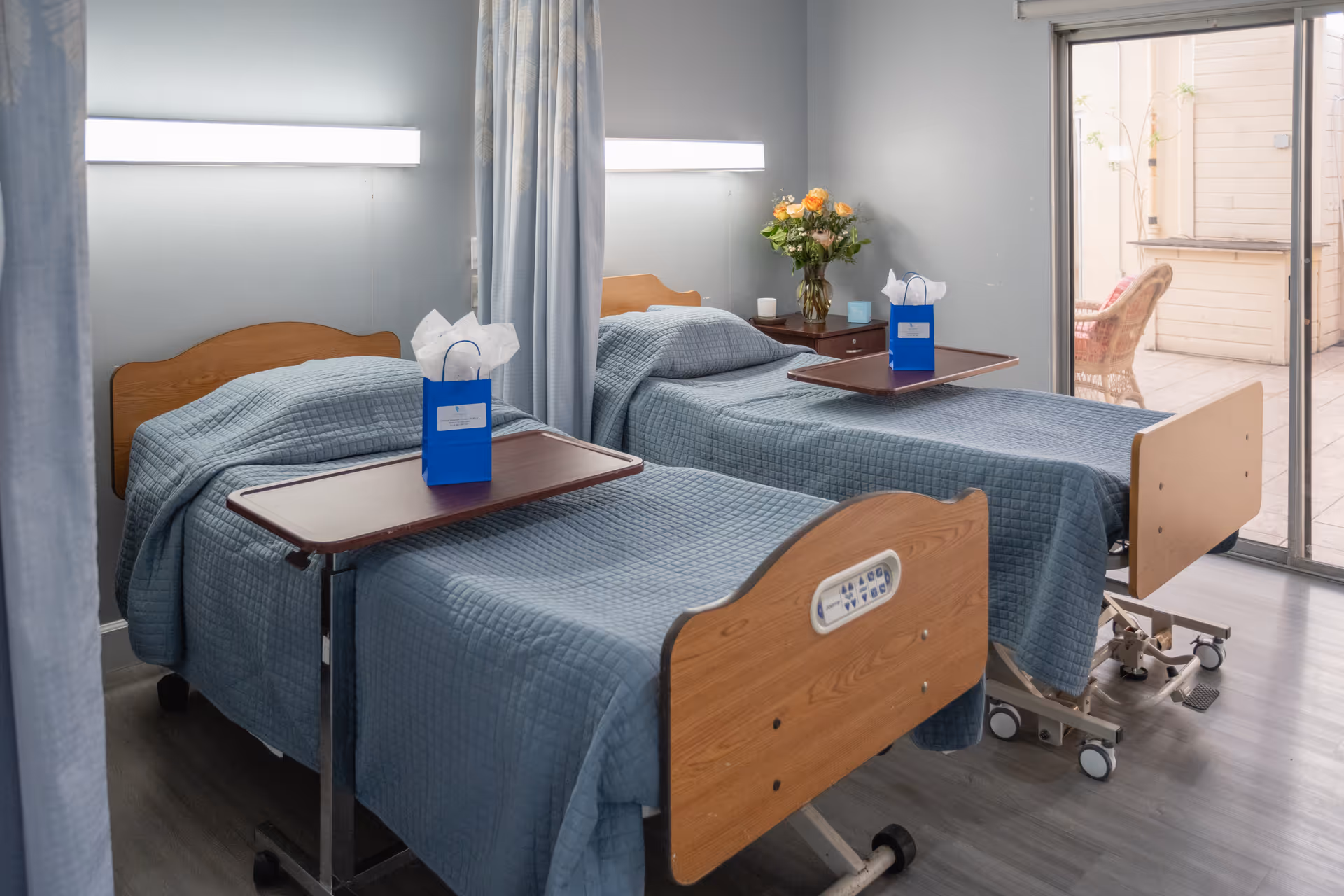 Two hospital-style beds with blue quilts, privacy curtains, and sliding glass door in a patient room.