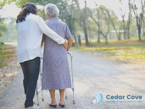 A younger woman walking outdoors on a path, supporting an elderly woman who is using a walker. They are seen from behind, surrounded by trees and greenery in a peaceful setting.