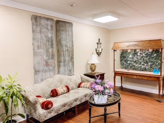 A cozy living room area with a vintage-style beige sofa adorned with red and beige cylindrical pillows. In front of the sofa is a round glass-top coffee table with a floral arrangement in a white vase. To the left of the sofa is a green leafy plant. Behind the sofa, there are three large rustic wooden panels mounted on the wall. To the right, there is a wooden console table with a decorative green plant wall behind it and two blue bottles on top. A table lamp with a white shade sits on a small side table next to the sofa. The room has wooden flooring and beige walls with a white ceiling and a square ceiling light.