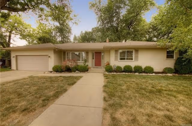Single-story ranch-style house with a red front door, attached garage, and a walkway across a front lawn surrounded by trees.