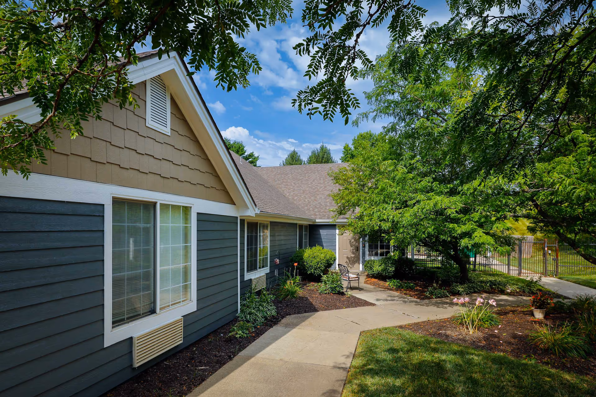 Exterior view of a single-story assisted living building with a walkway, landscaping, and trees.
